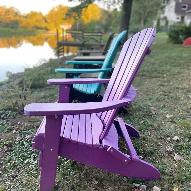 A row of purple and teal adirondack chairs are sitting next to a body of water.