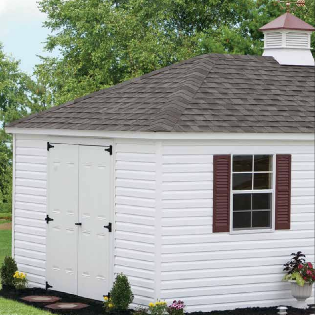 Tan shed with three windows, oval window, and shingled roof, set against trees.