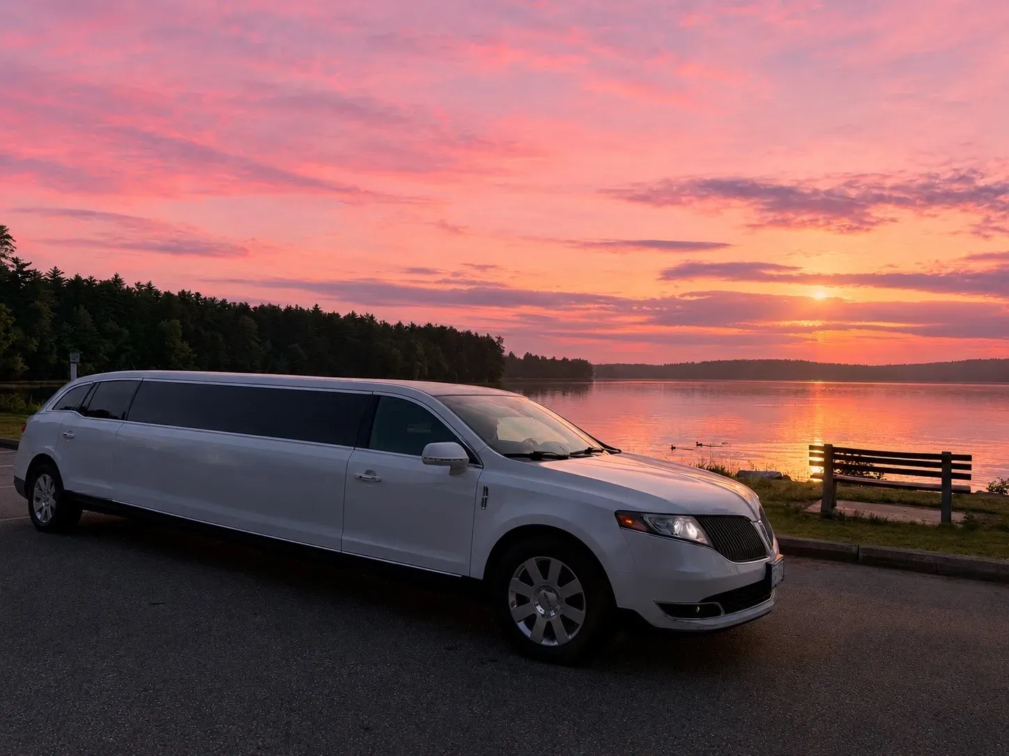 White stretch limousine parked by a lake at sunset with pink-orange sky and trees in the background