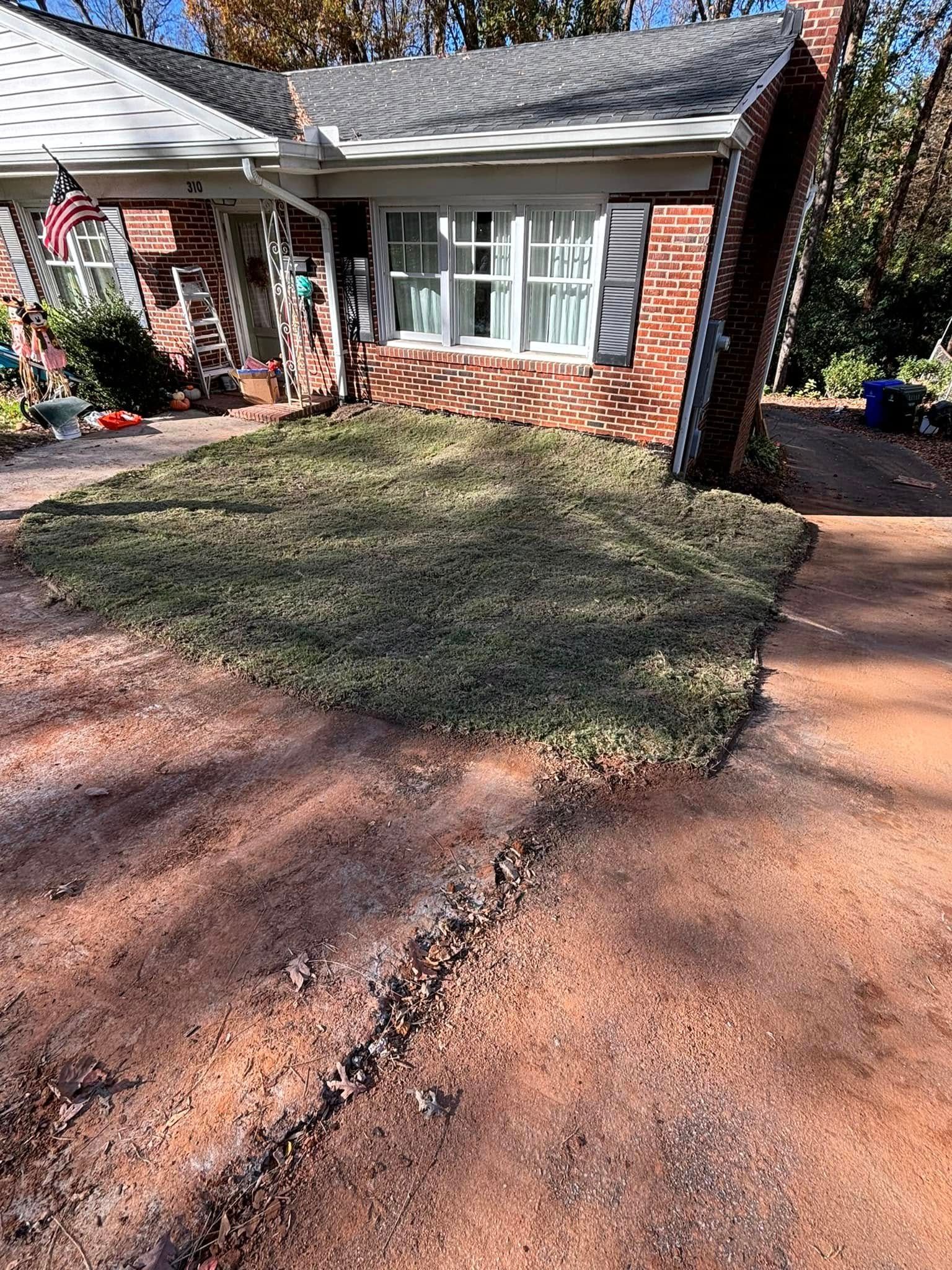 House with a freshly laid patch of sod in front. Brown driveway, red brick facade, and a flagpole visible.