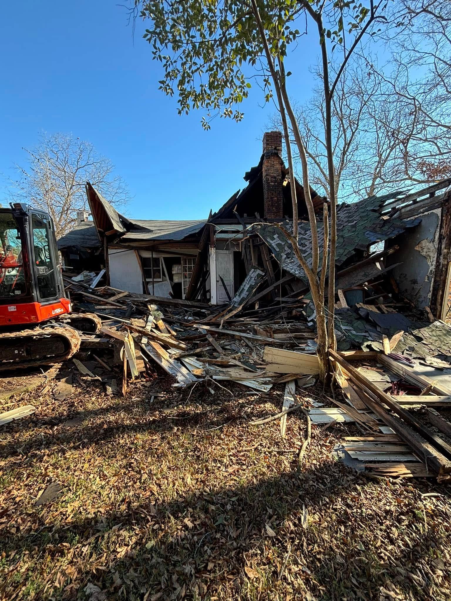 A severely damaged house undergoing demolition. Debris litters the ground, with an excavator visible on the left.