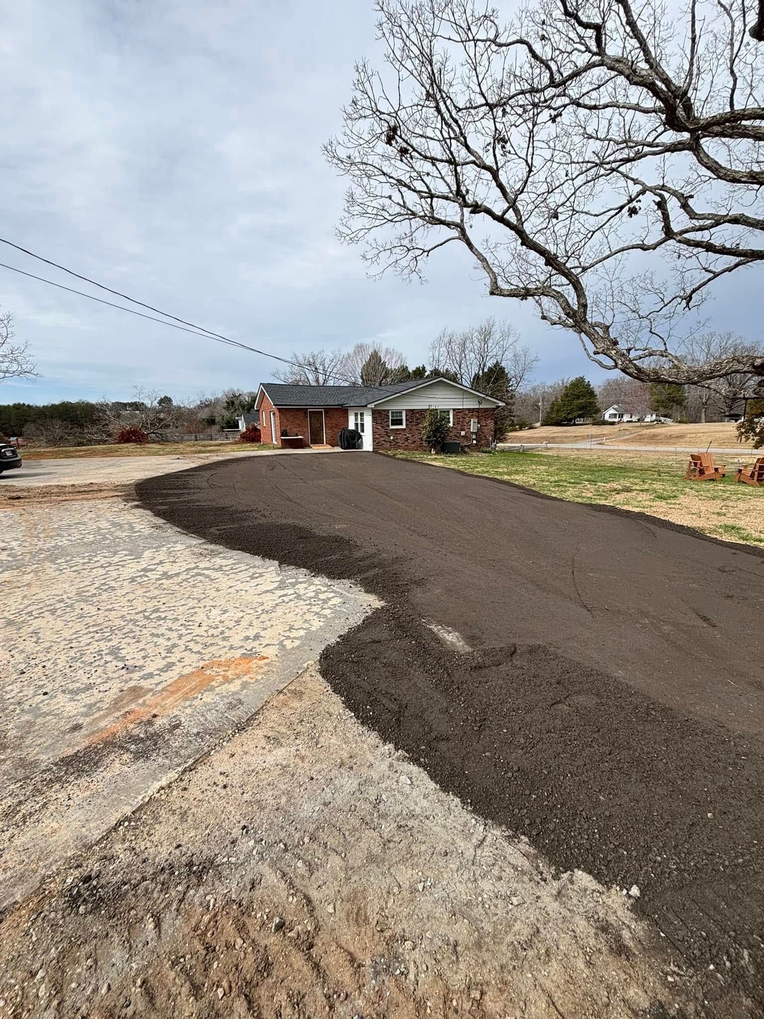 A dirt driveway being graded in front of a brick house on a cloudy day.