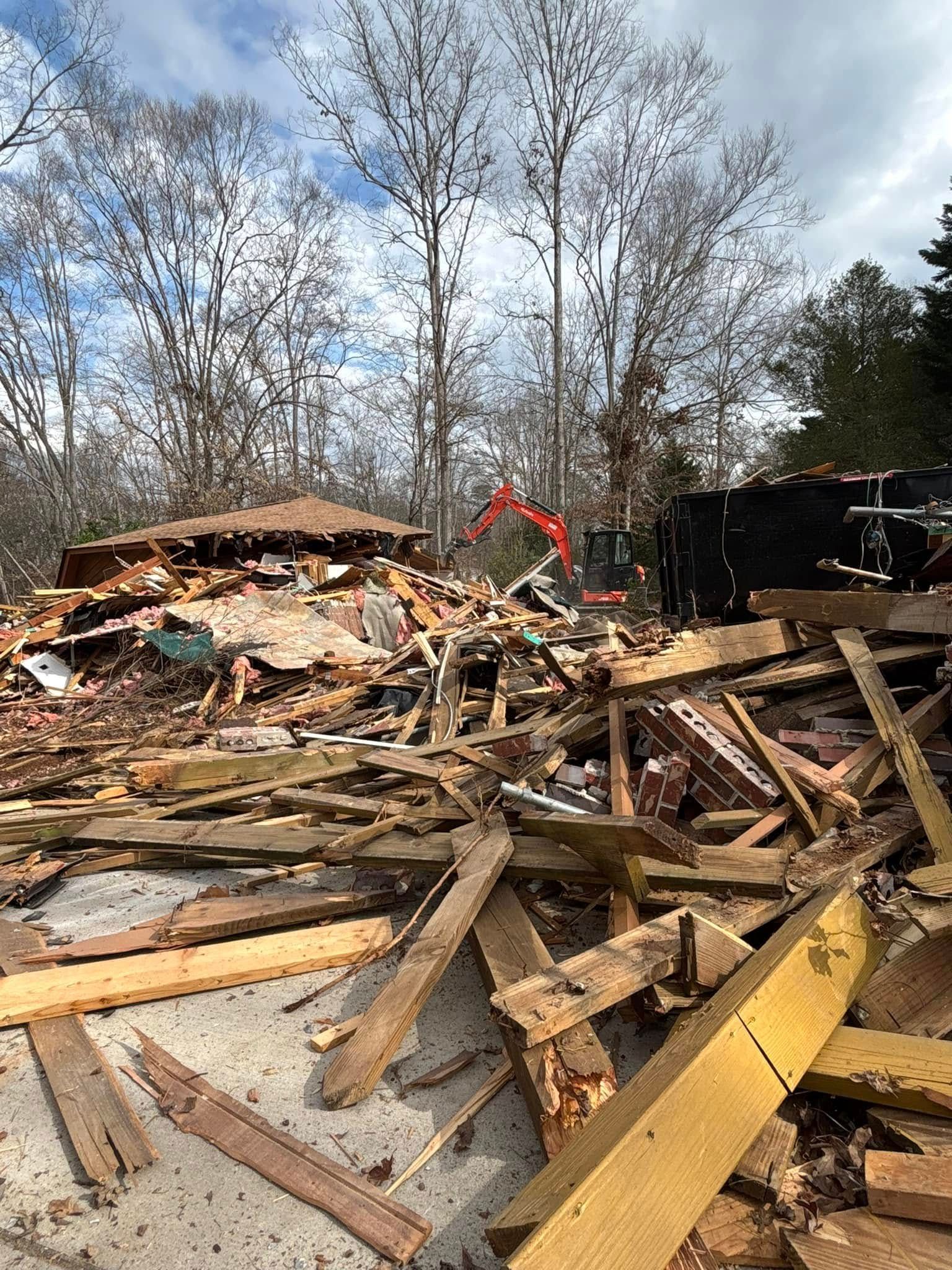 A building being demolished, with debris and an excavator. The sky is cloudy, and trees are in the background.