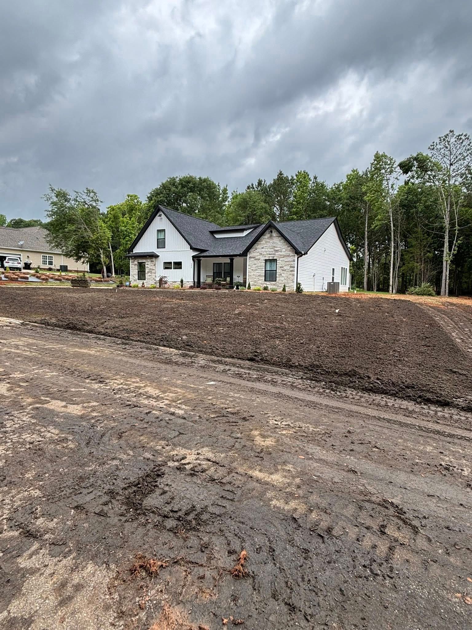 A modern house under construction with a black roof, white siding, and stone accents. The yard is muddy.