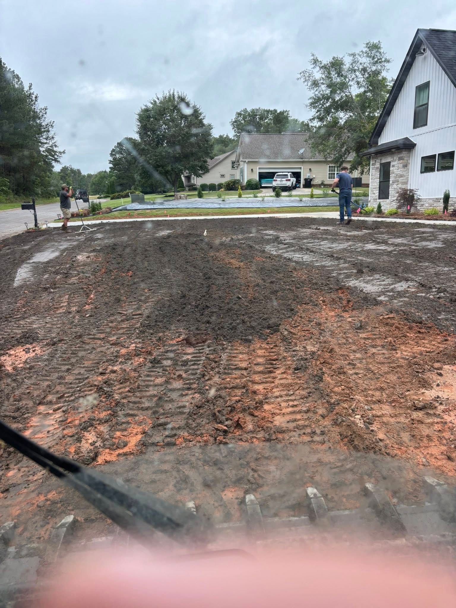 A front yard being graded. The soil is brown and churned. A person stands near a white house under a cloudy sky.