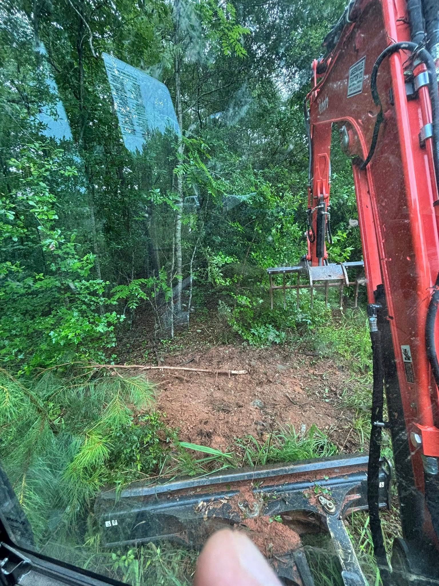 View from inside an excavator looking out at a clearing in a green wooded area.
