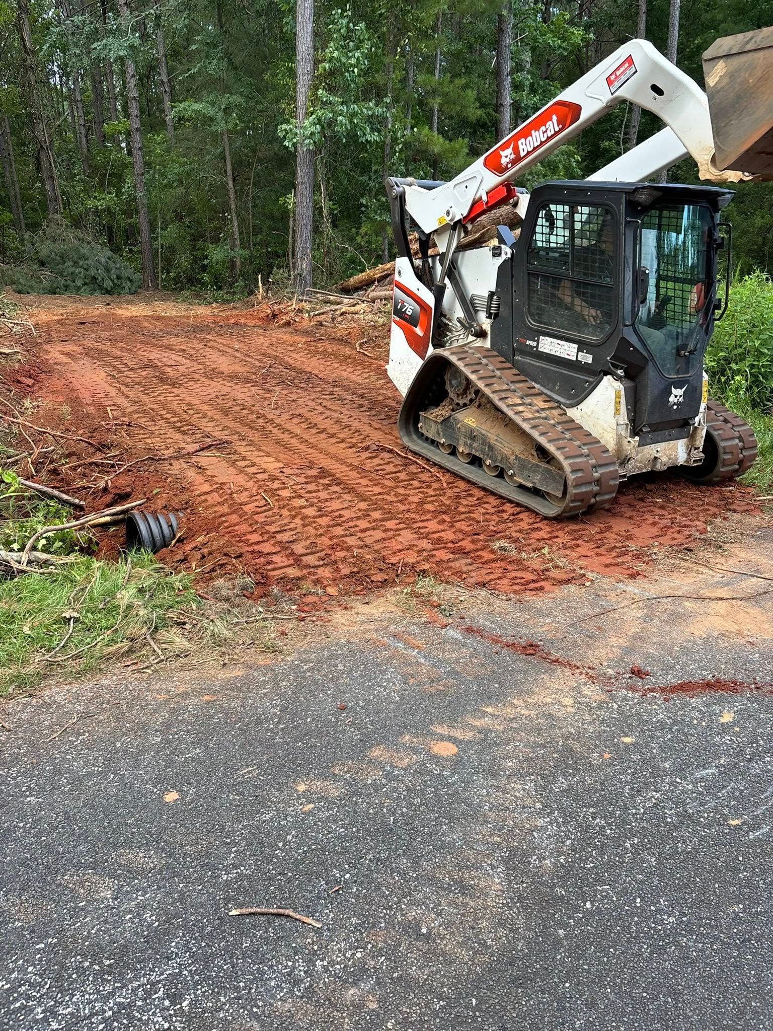 A Bobcat skid-steer loader on a red-dirt construction site, near trees, parked in front of an asphalt area.