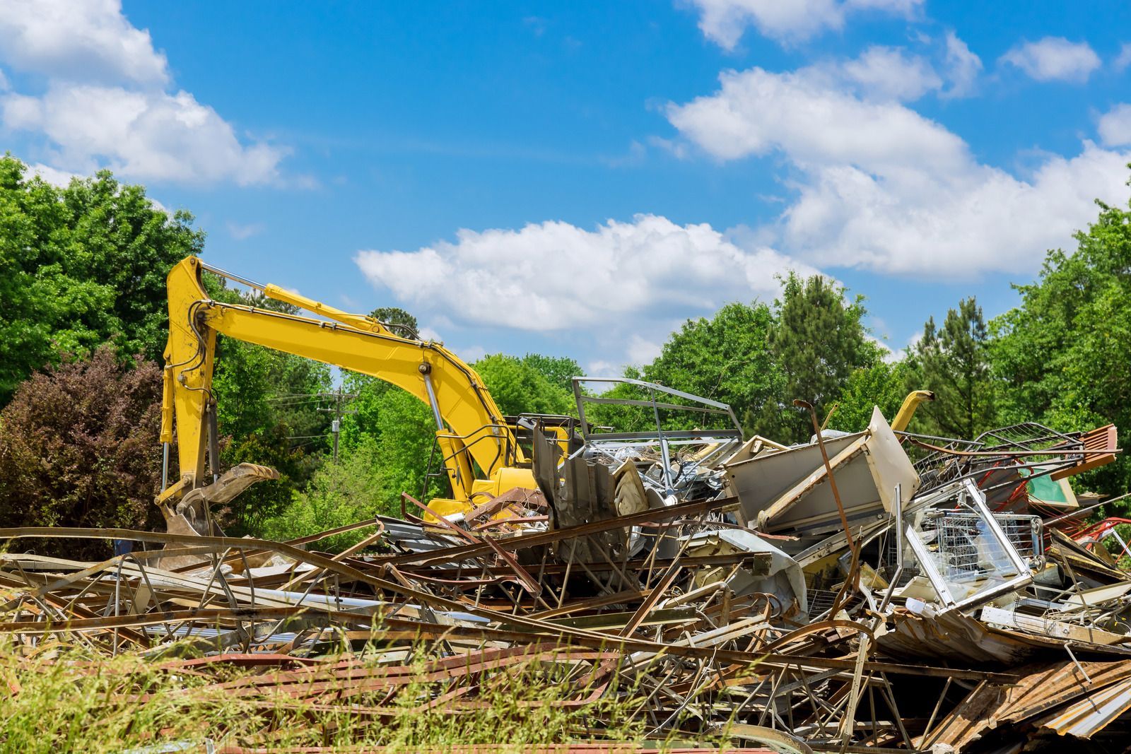 A small brick house with a chimney being demolished by an excavator; trees surround the house.