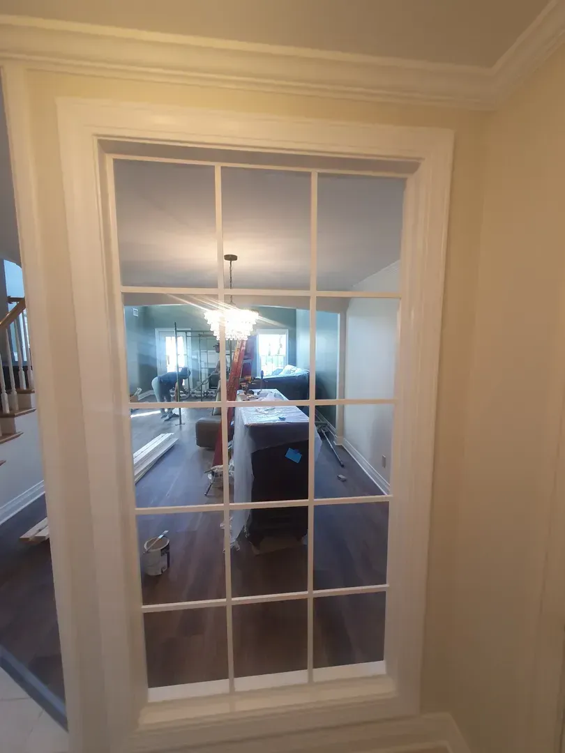 View through a white-paned interior window into a warmly lit hallway with a black dresser and staircase.