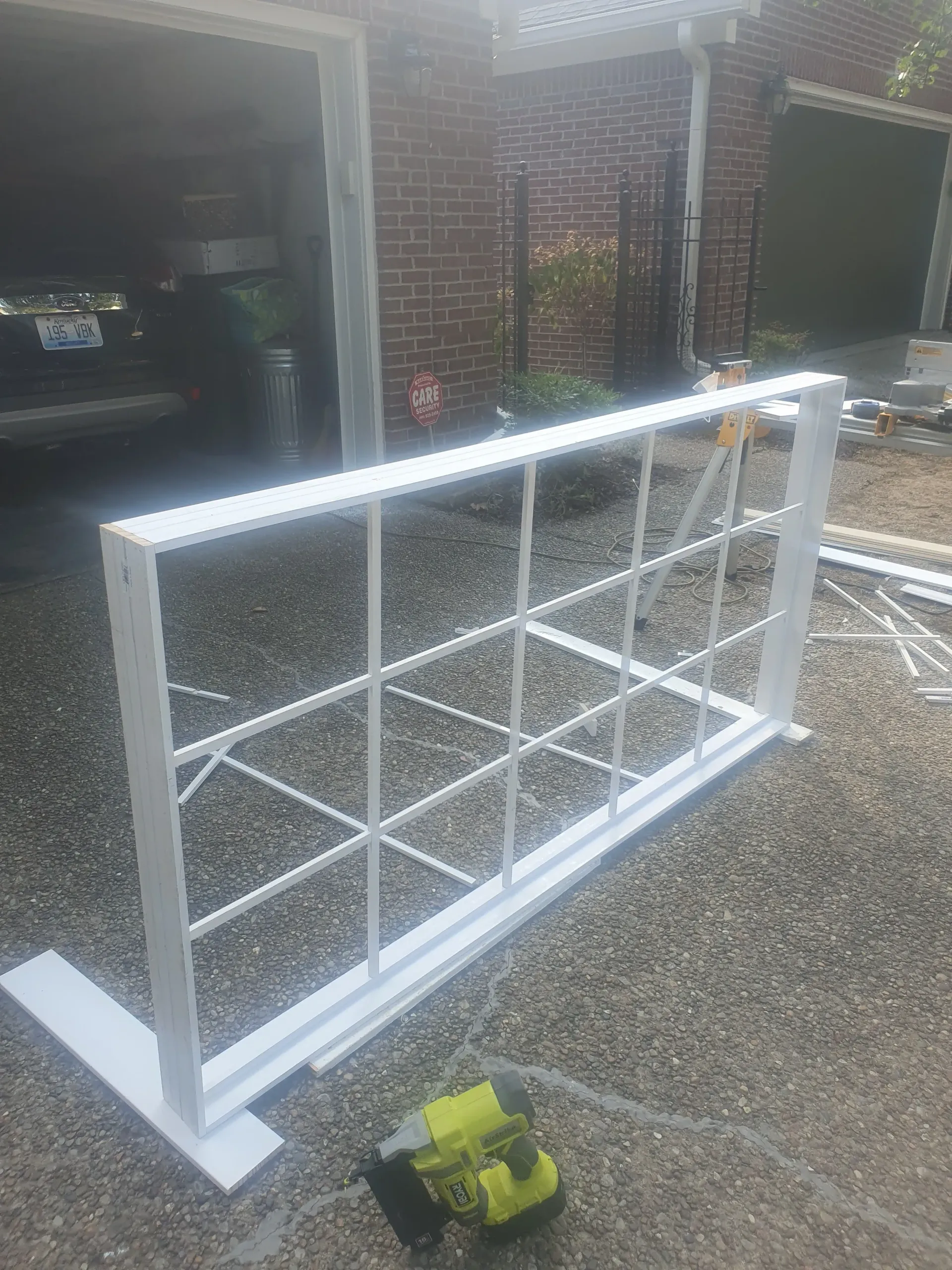 White metal frame with wire mesh leaning on driveway, with a yellow tape measure in front of a brick house.