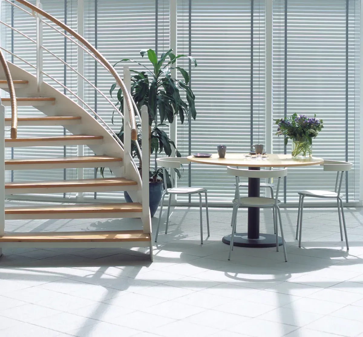 Bright modern atrium with a curved staircase, round table, chairs, and potted plants by large windows