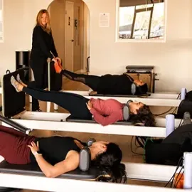 People lying on Pilates reformers in a studio while an instructor stands nearby