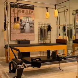 Pilates reformer machine with yellow bench in a studio, hanging straps, and wall poster.