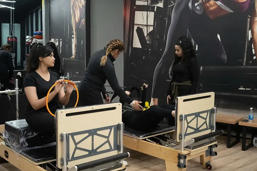 Three people exercising in a gym, using resistance bands and reformer machines in front of a mirrored wall.