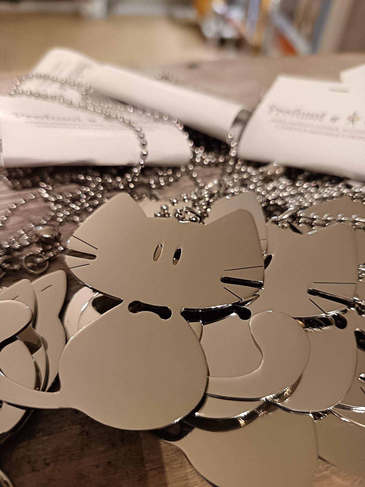 Ciondoli in argento a forma di gatto su catene a palline, con targhette sullo sfondo.