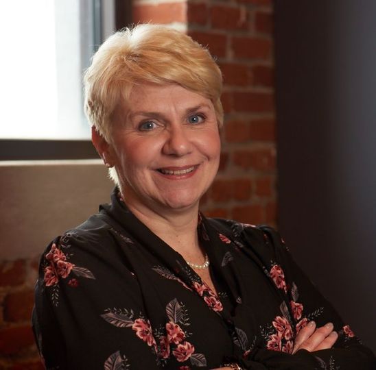 Smiling person with short blond hair in a black floral blouse, standing by a window and brick wall.