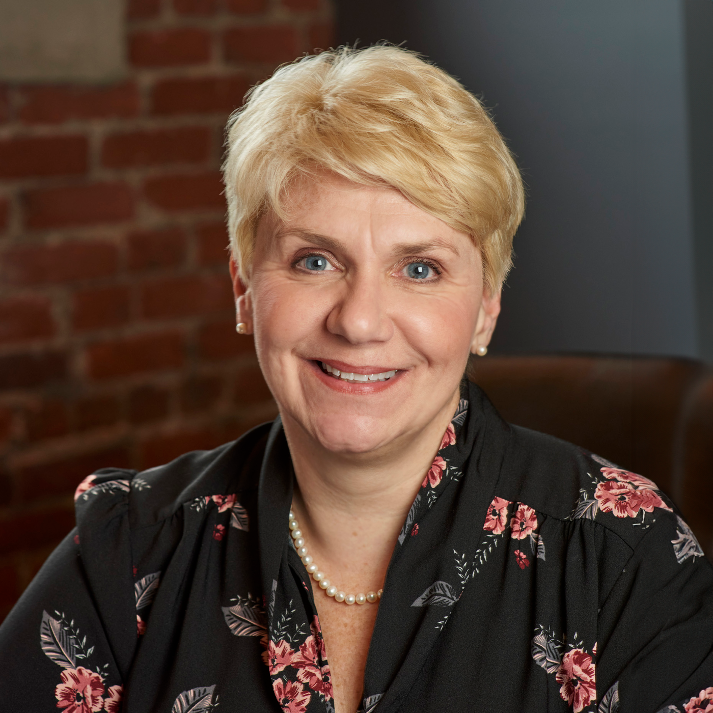 Smiling woman with short blonde hair in a black floral blouse, seated indoors against a brick wall.
