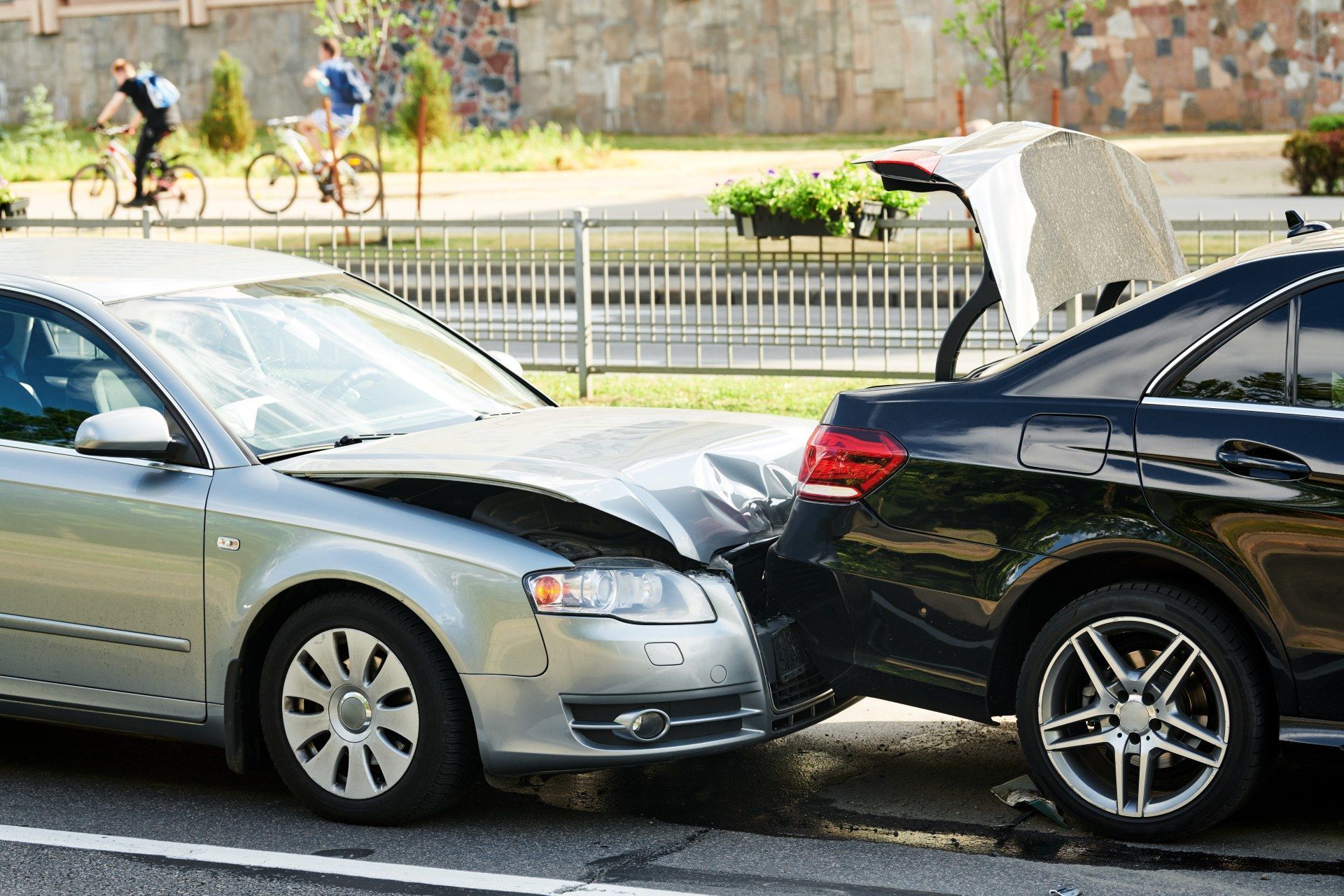 Silver car rear-ending black sedan on a street, with bikes and pedestrians in the background.