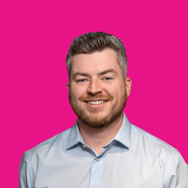 Man with a beard smiling, wearing a light blue button-up shirt, set against a pink background.