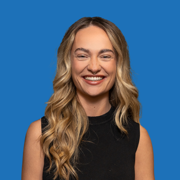 Woman with long wavy blonde hair smiling, wearing a black top, against a blue background.