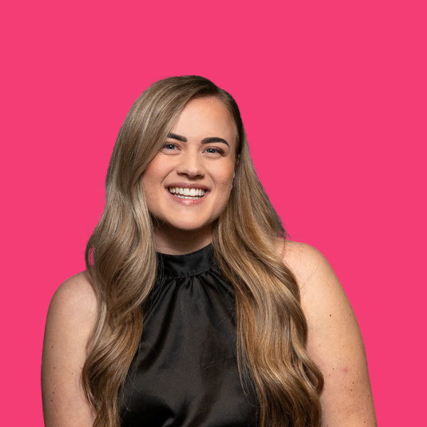 Woman with long wavy blonde hair, smiling, wearing a black top, against a pink backdrop.