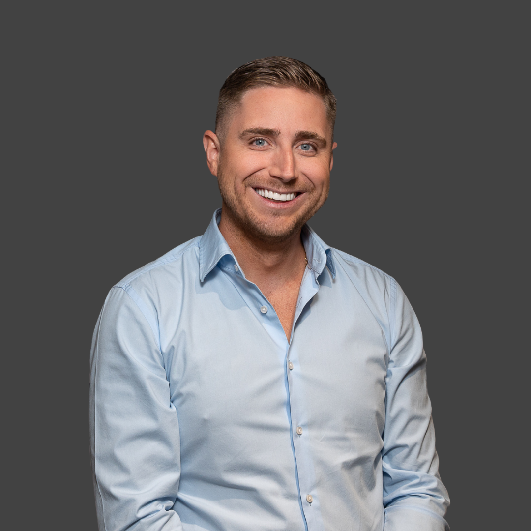 Man in a light blue button-down shirt smiles broadly, set against a gray background.
