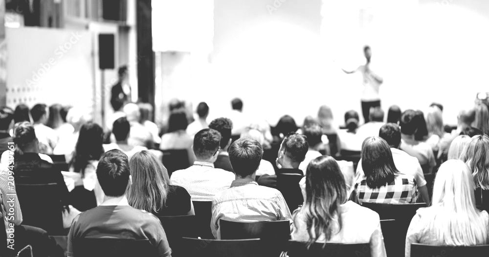 A large group of people are sitting in a lecture hall watching a presentation.