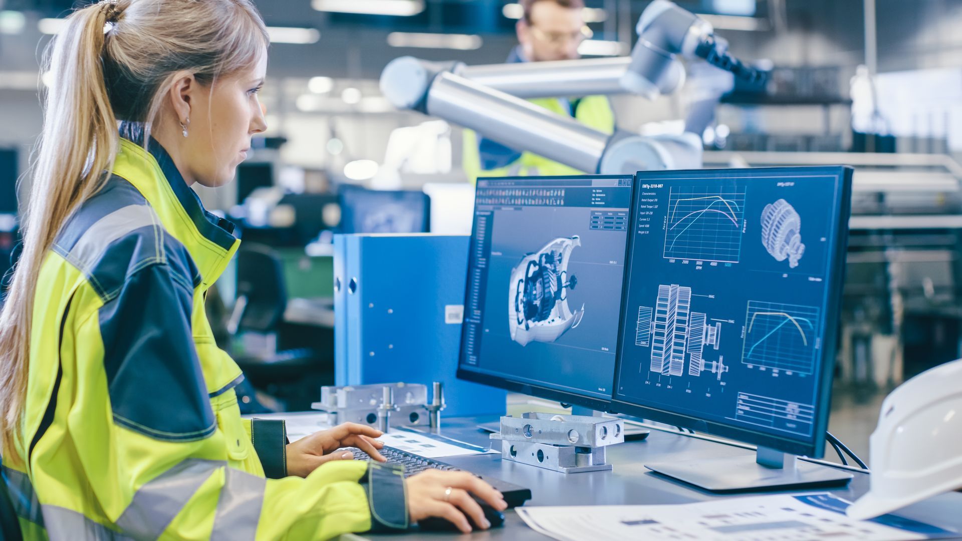 A woman is sitting at a desk in front of a computer in a factory.