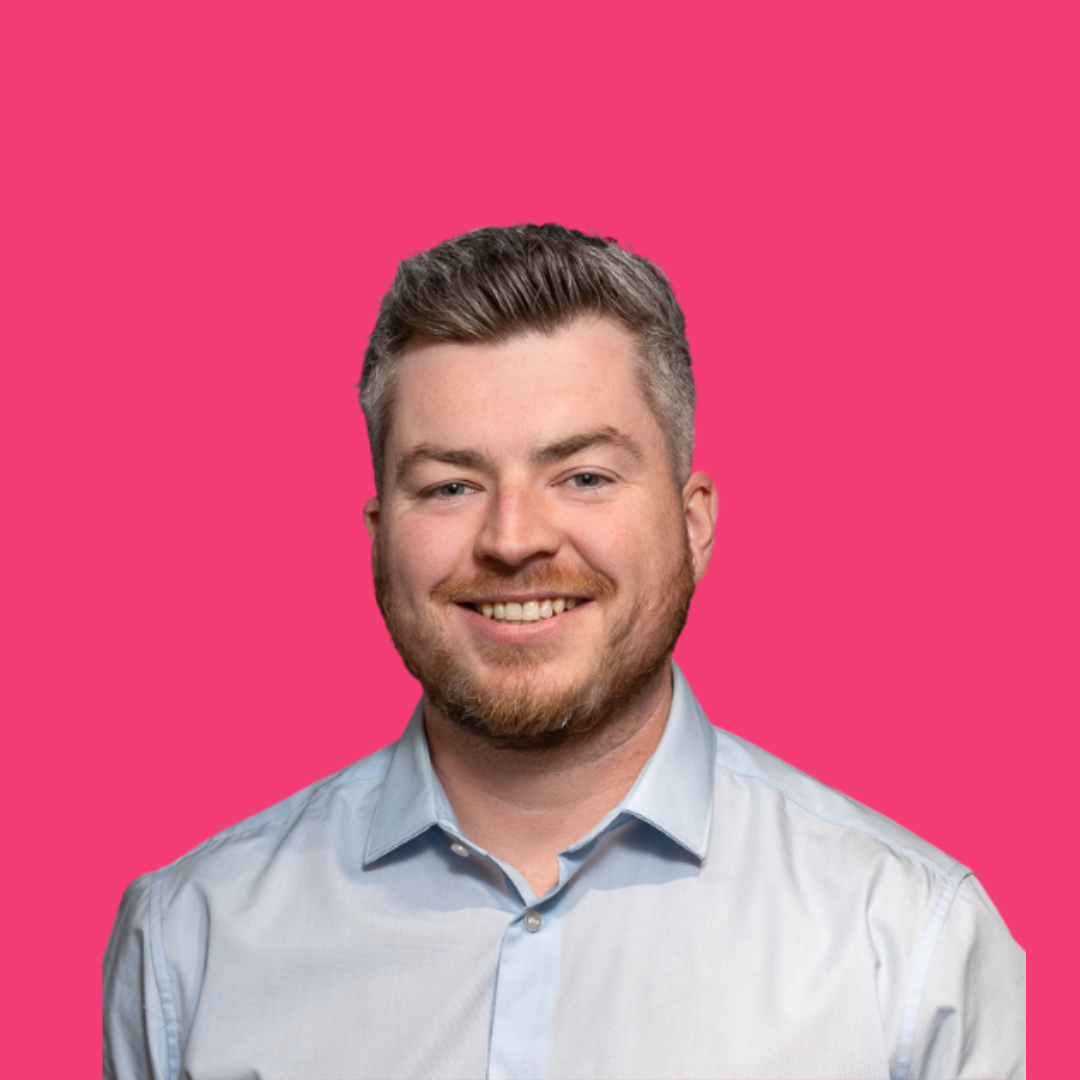 Man with short, gray hair and a beard, smiling. Wearing a light blue button-down shirt against a pink background.