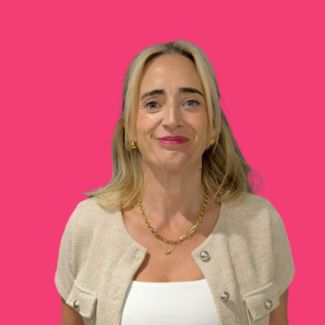 Woman with blonde hair wearing a beige sweater and gold jewelry, standing against a pink background.