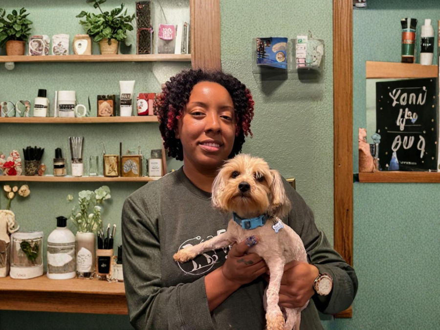 Person holding a small tan dog in a shop with wooden shelves of plants and candles behind them