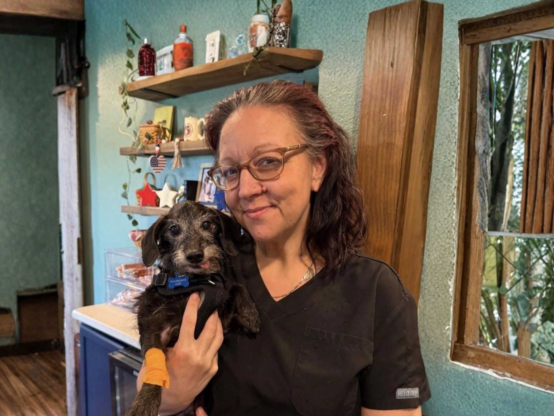 Woman holding a small black dog indoors beside a wooden shelf and window