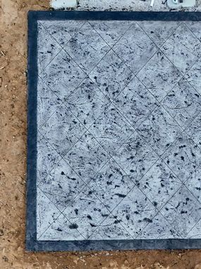 A close up of a gray tile with a black border on a dirt surface.