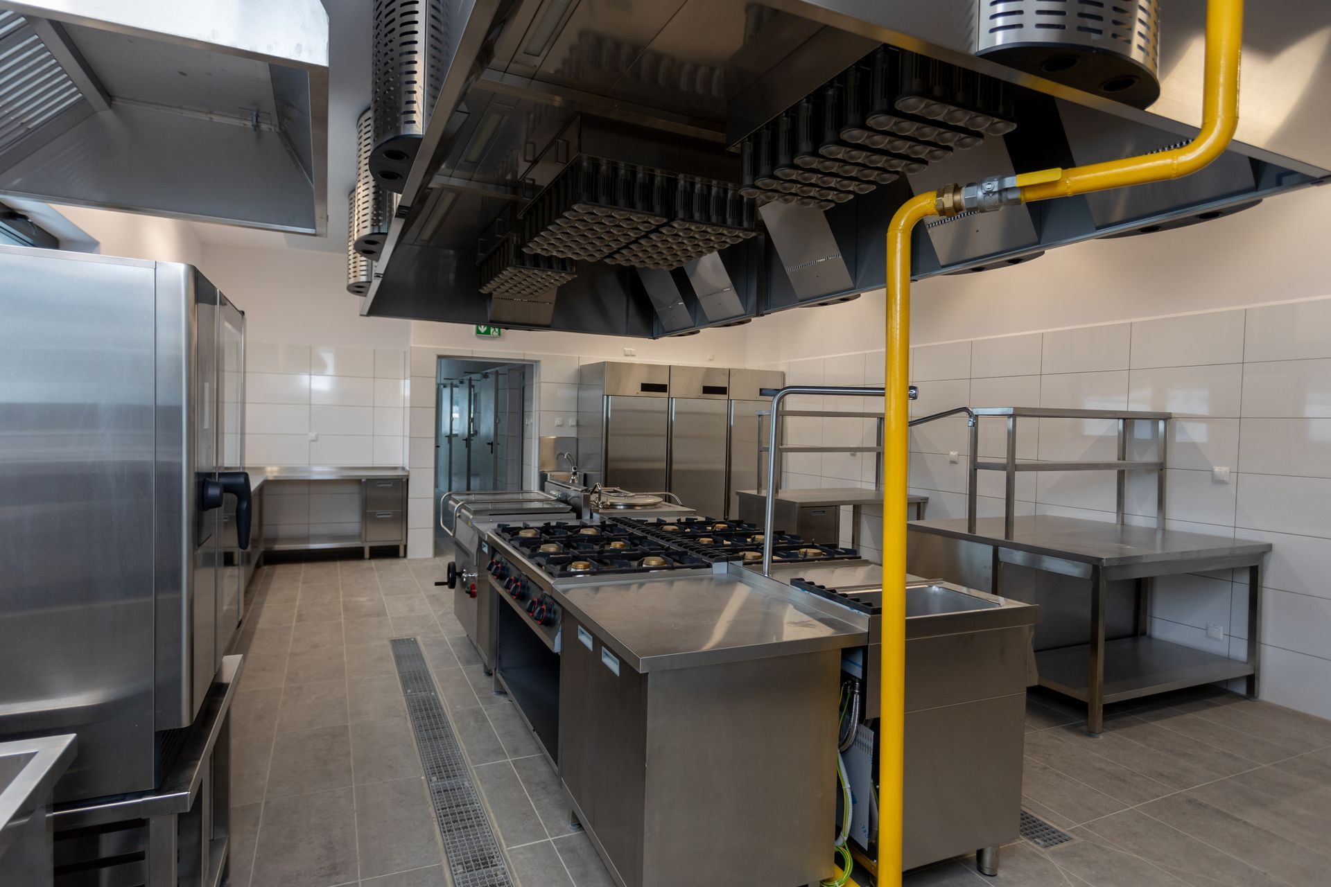 Industrial kitchen with stainless steel cooking equipment, tiled walls, and yellow gas pipes above. Industrial kitchen with stainless steel cooking equipment, tiled walls, and yellow gas pipes above.
