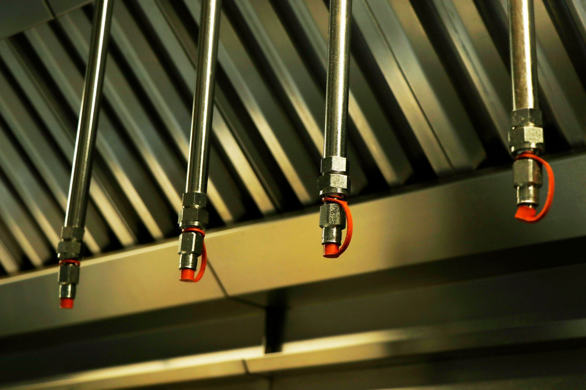 Five hanging industrial spray nozzles under a metal hood, with red tips and brass fittings. Five hanging industrial spray nozzles under a metal hood, with red tips and brass fittings.