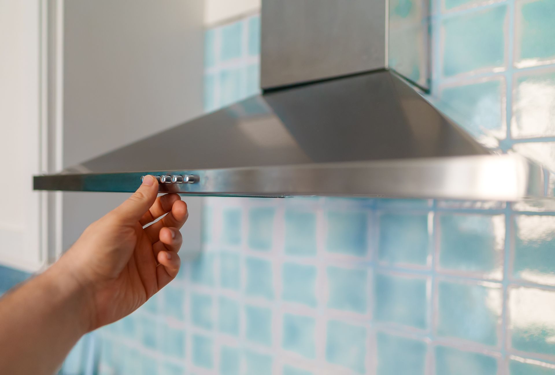 Hand adjusting a stainless steel kitchen range hood control against a blue tiled wall Hand adjusting a stainless steel kitchen range hood control against a blue tiled wall