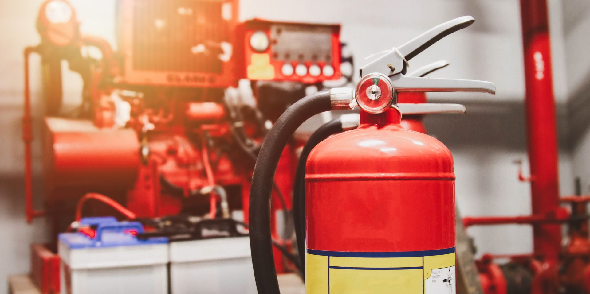 Red fire extinguisher in the foreground, with blurred industrial equipment in the background