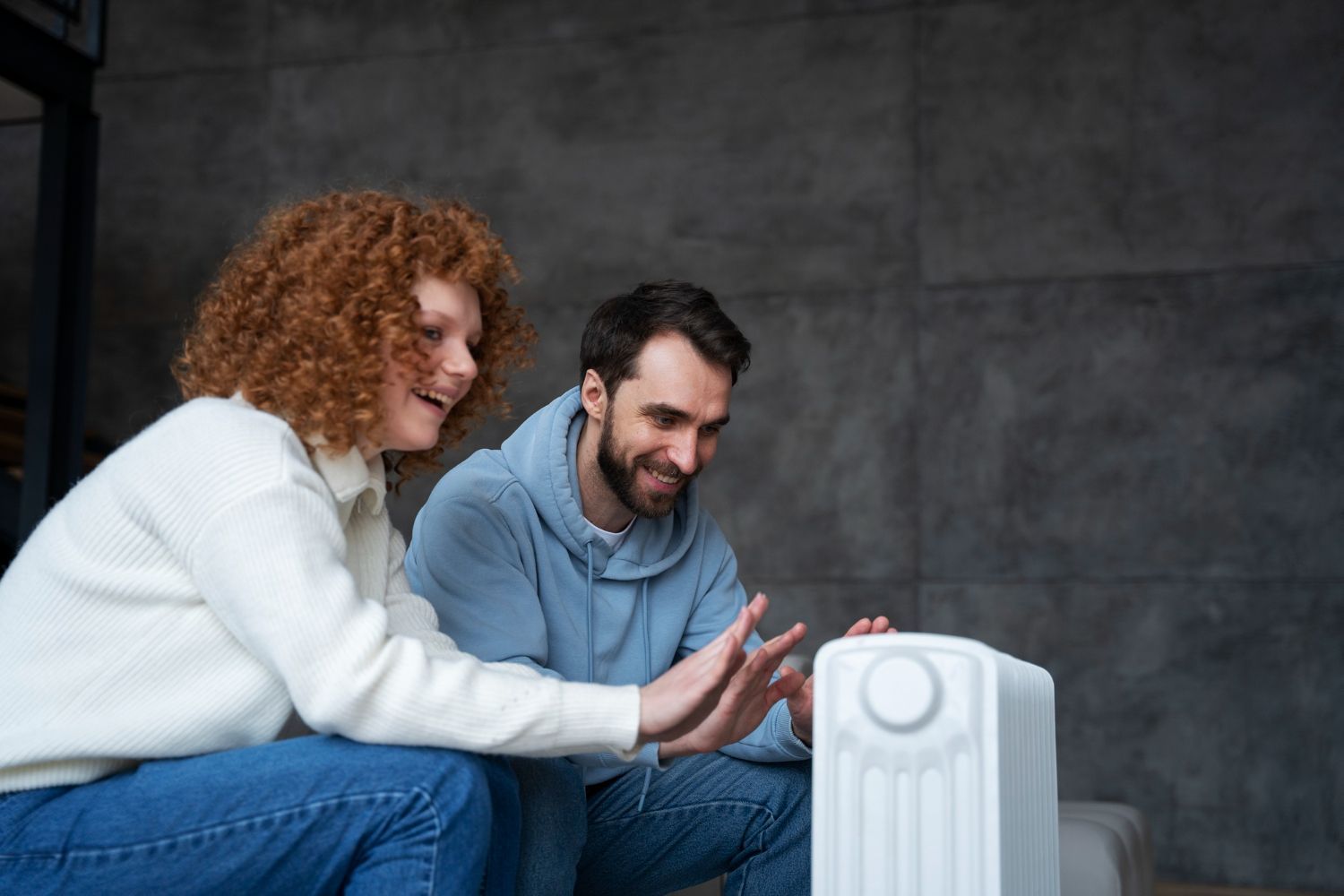 Woman and Man Sit Near a White Heater, Touching It With Their Hands — Vantage Plumbing and Drainage In Pacific Pines, QLD