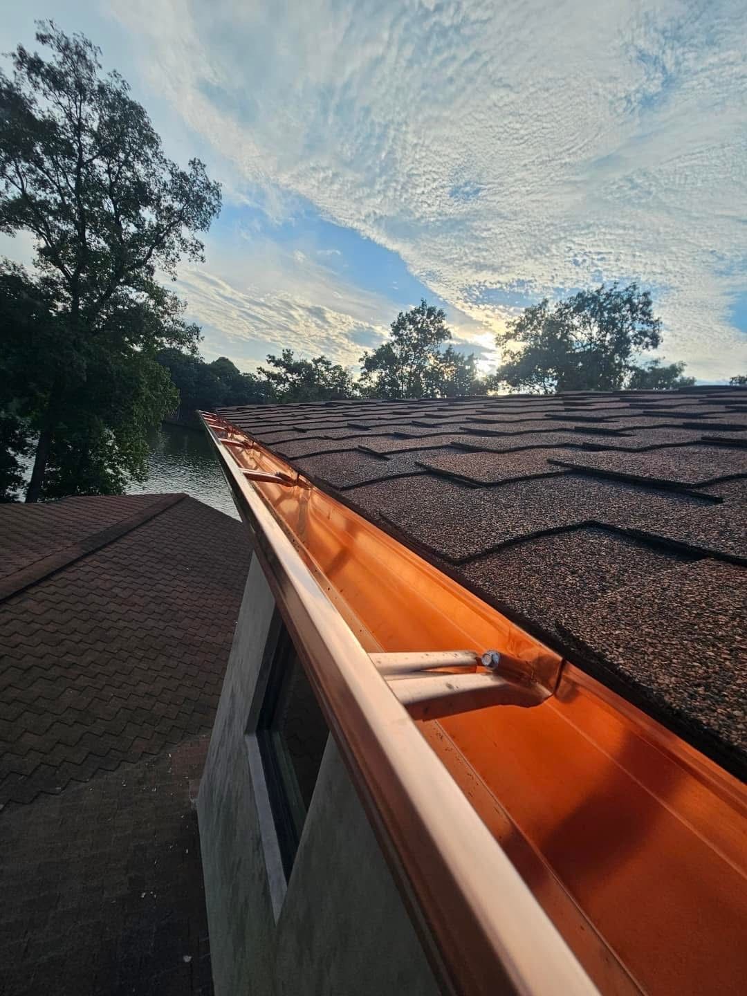 Copper gutters on a shingled roof under a cloudy sky, near trees.