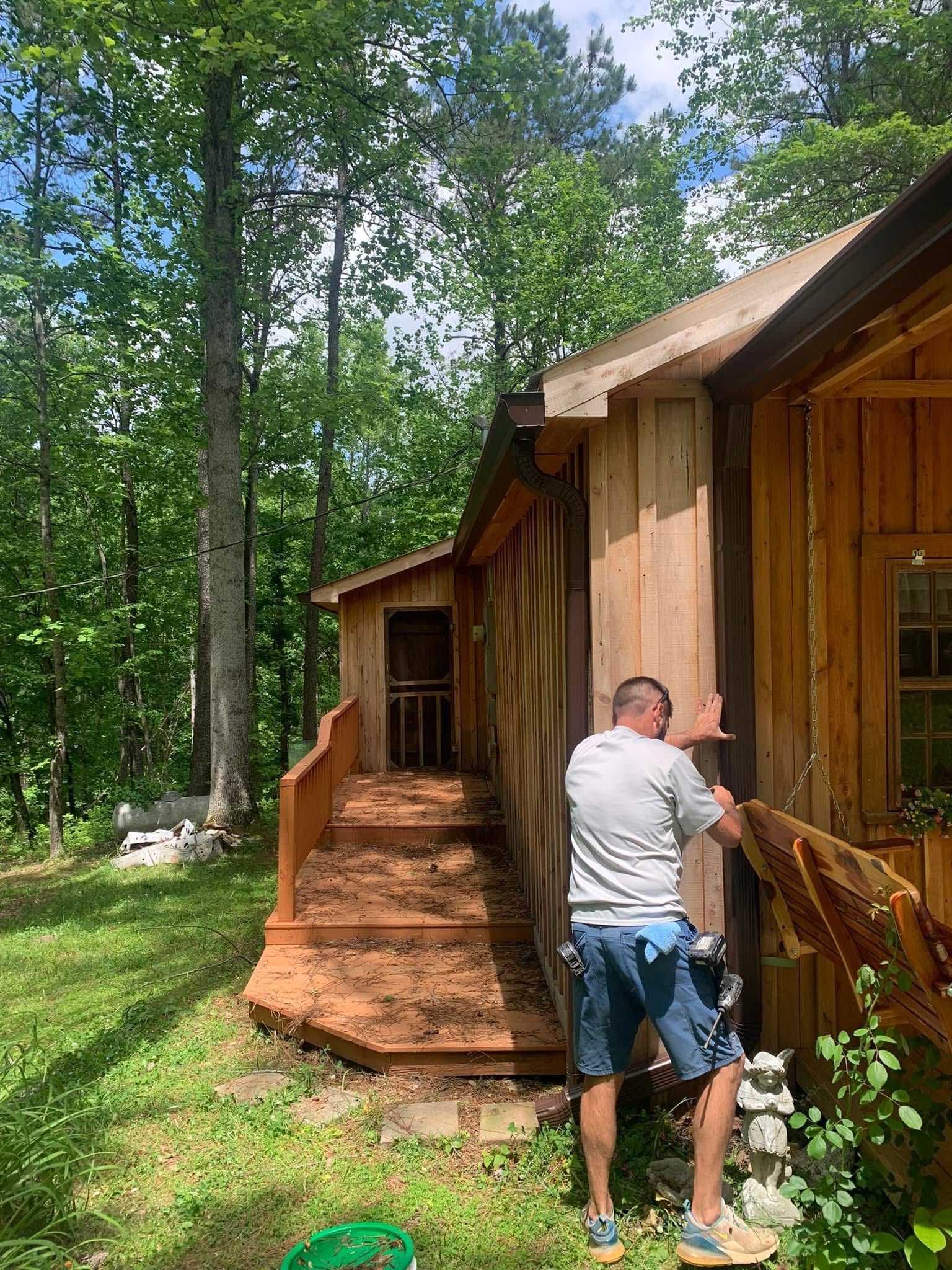 Man repairs wooden cabin siding, standing on a porch with stairs. Forest background.