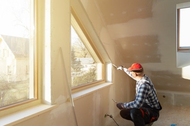 Worker in red hard hat applying drywall compound to a sloped wall near windows