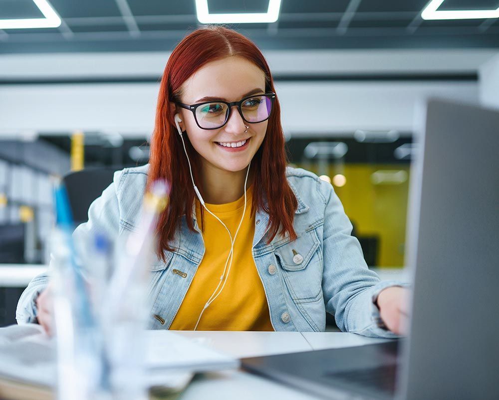 A woman wearing glasses and headphones is sitting at a desk in front of a laptop computer.