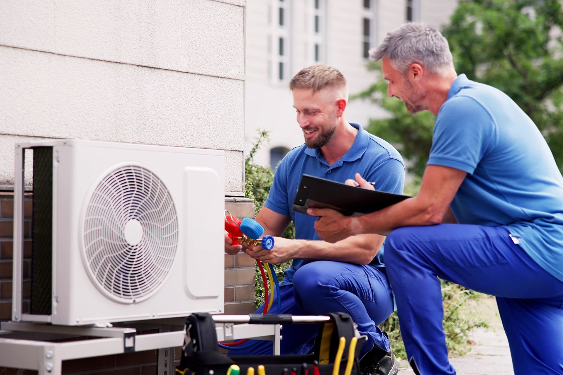 Two HVAC technicians working on an outdoor air conditioning unit. One kneels and works with gauges. The other consults a clipboard.