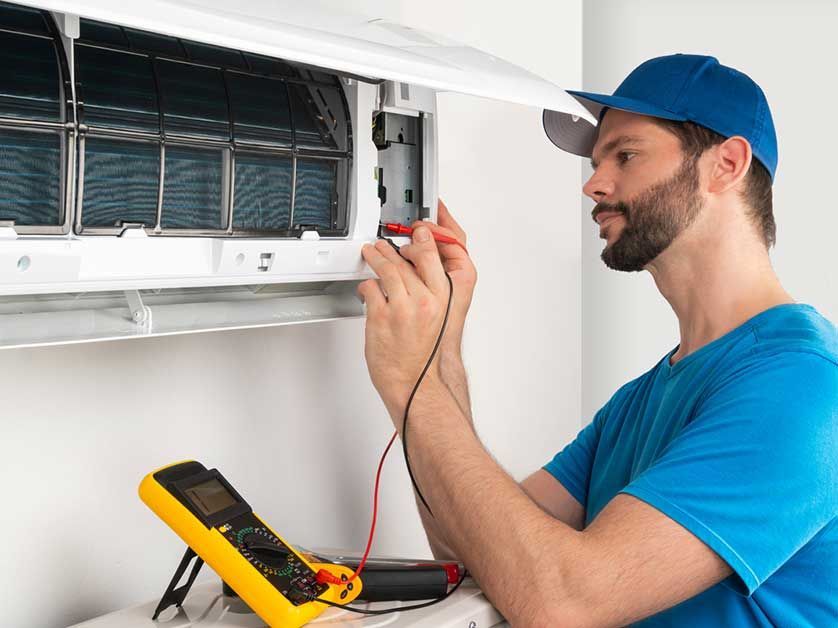 Man in blue shirt and cap uses a multimeter on an air conditioner indoors.