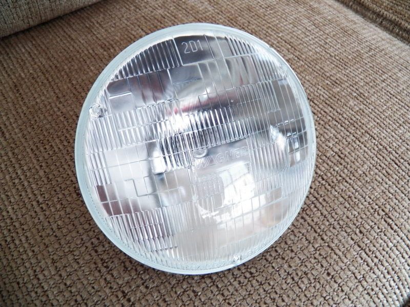 A round, clear headlight with a textured glass surface, resting on a brown, textured surface.