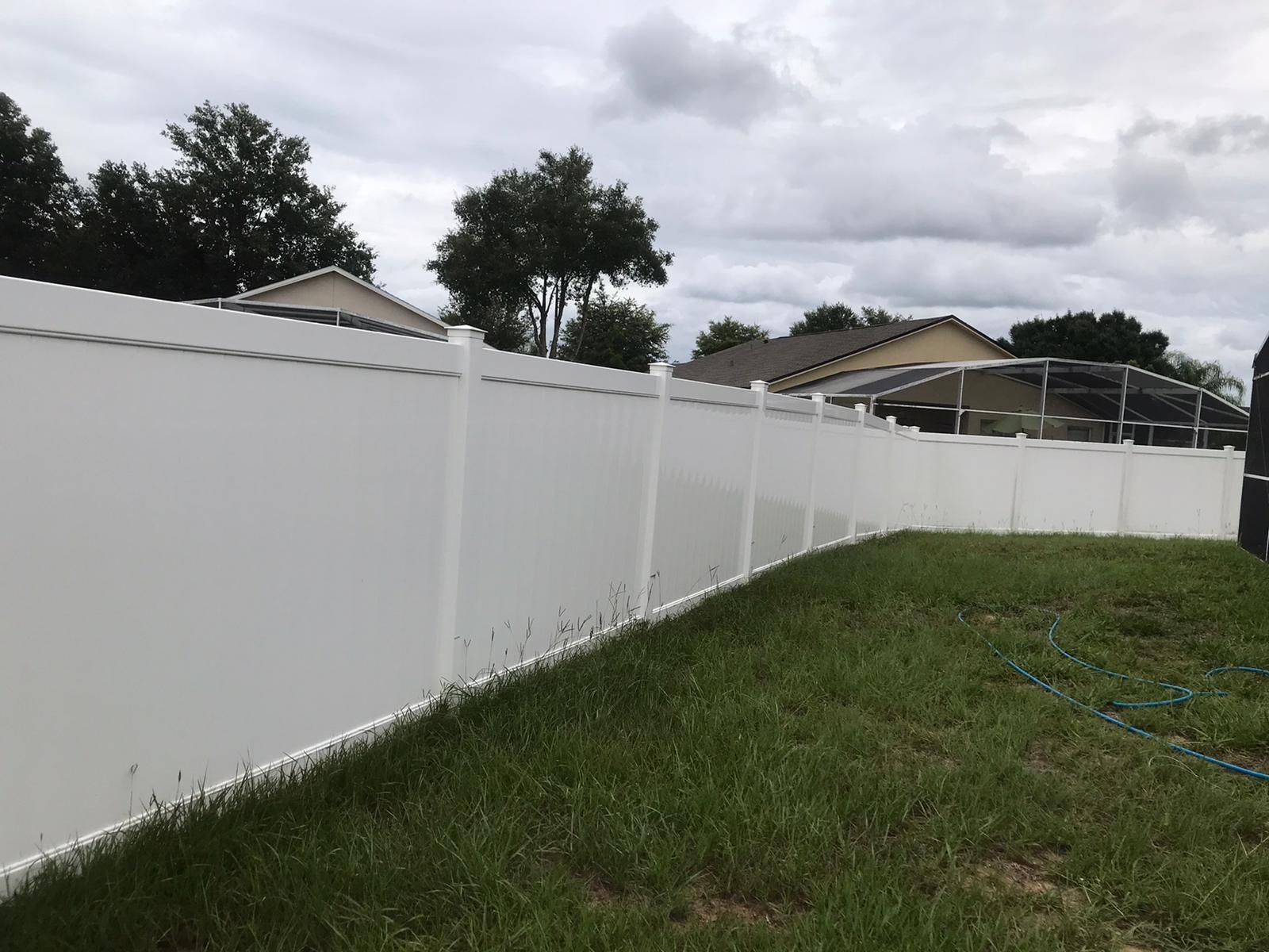 White vinyl fence along a grassy yard under cloudy skies with trees and rooftops in the background