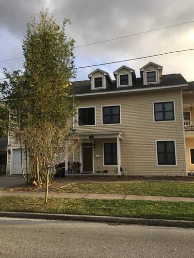 Two-story beige house with dormer windows, small porch, and a tree in the front yard beside a street