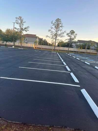 Empty paved parking lot with white lines at dusk, bordered by trees and a small building in the background