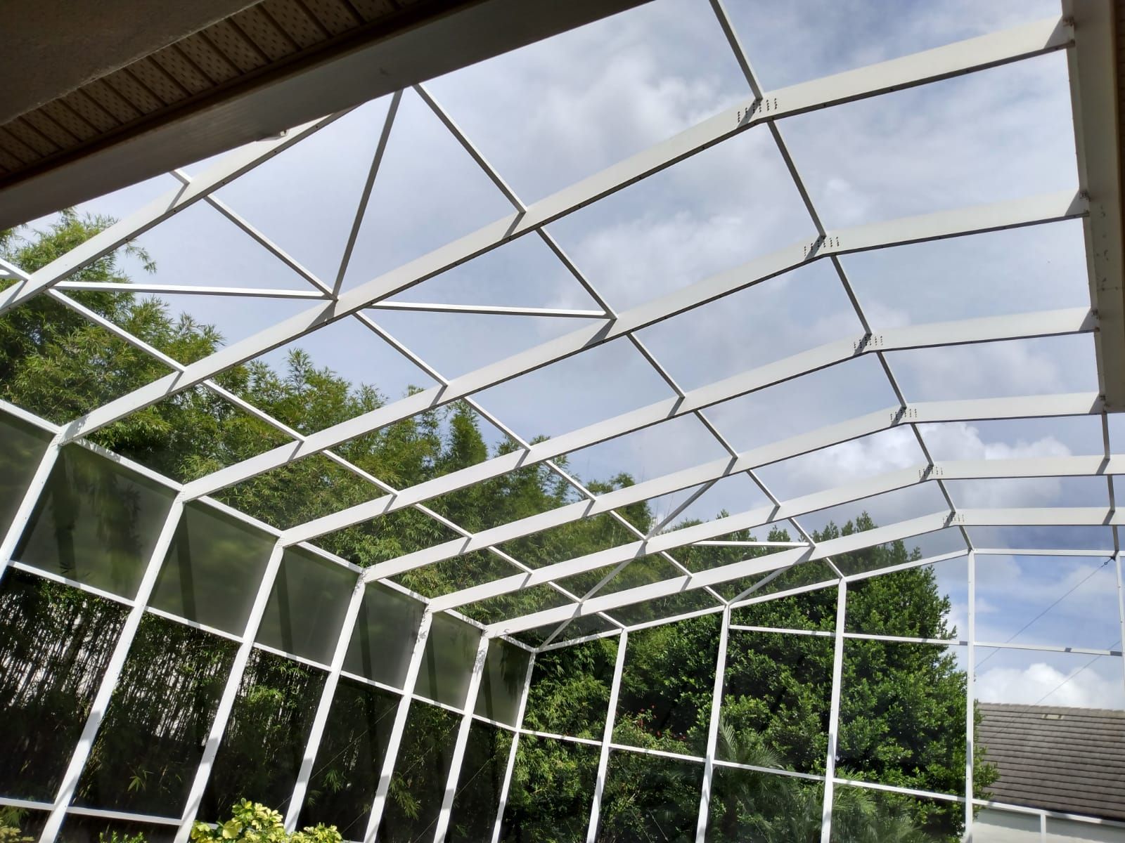 White metal patio frame with a translucent roof over green trees and a cloudy sky