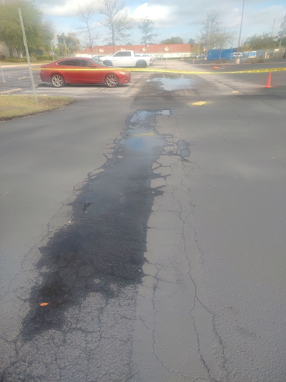 Cracked parking lot pavement with a long dark asphalt patch and a red car in the background.